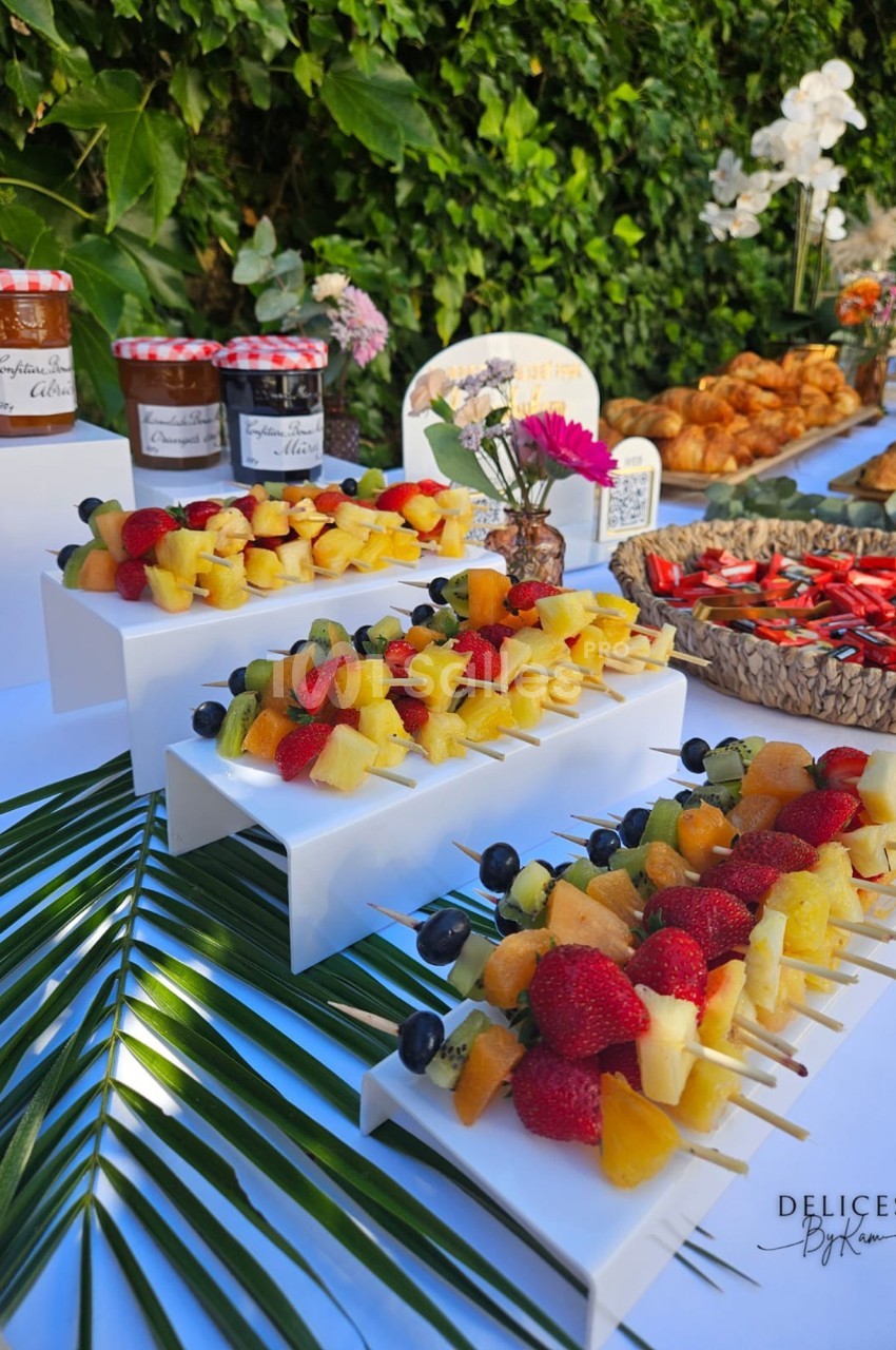 Brochettes de fruits frais disposées sur des supports blancs, avec confitures et viennoiseries en arrière-plan.