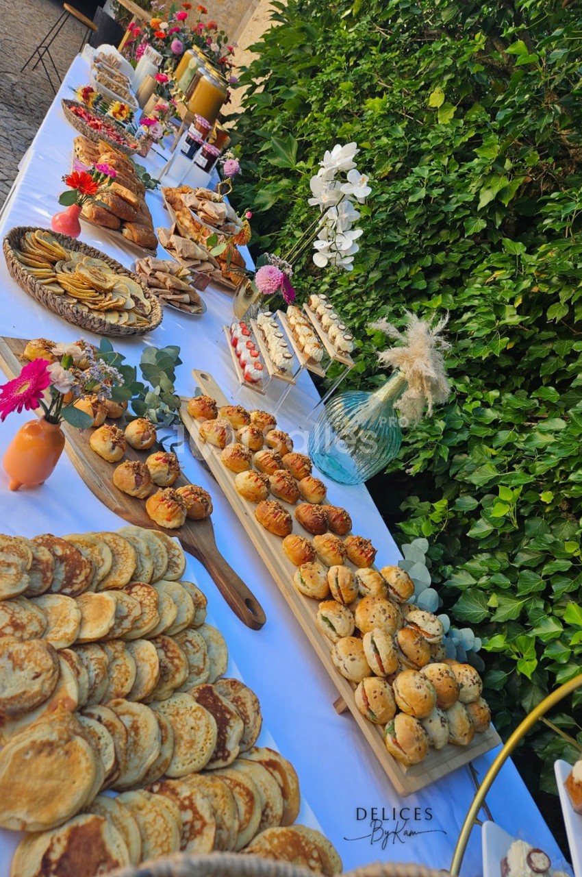Table garnie de pâtisseries variées, crêpes, muffins et décorations florales, disposée en extérieur près d'un mur végétal.