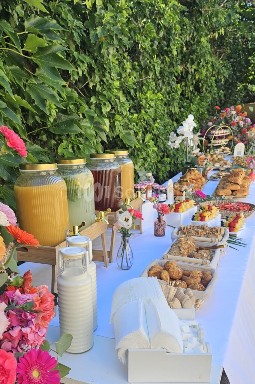 Buffet en extérieur avec boissons, viennoiseries, fruits et fleurs colorées sur une table blanche près d'un mur végétal.