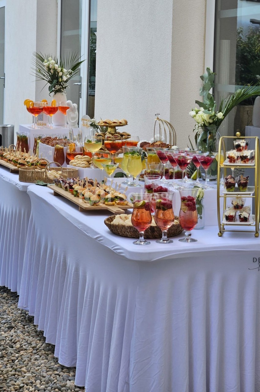 Buffet élégant en extérieur avec boissons, verrines, pâtisseries et décorations florales sur nappes blanches.