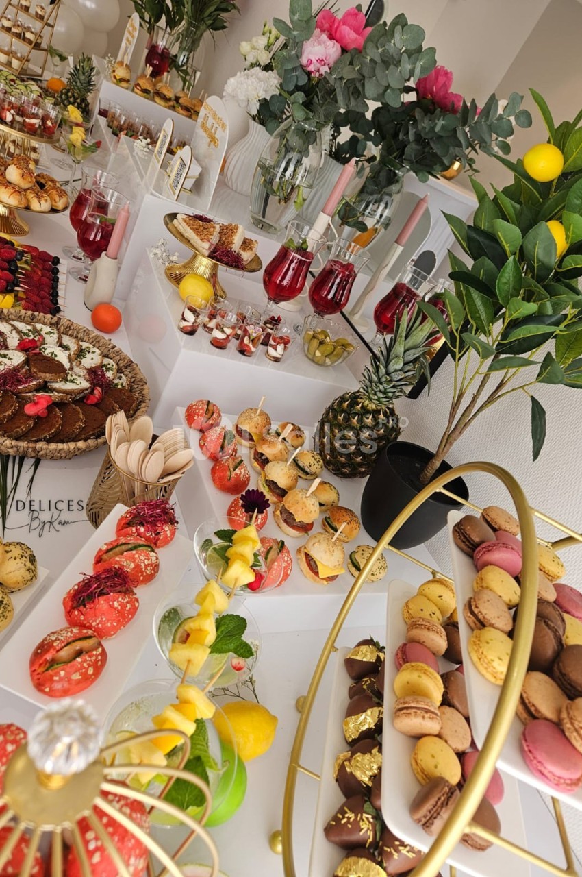 Buffet coloré avec macarons, fruits, pâtisseries et boissons, décoré de fleurs et de plantes vertes.