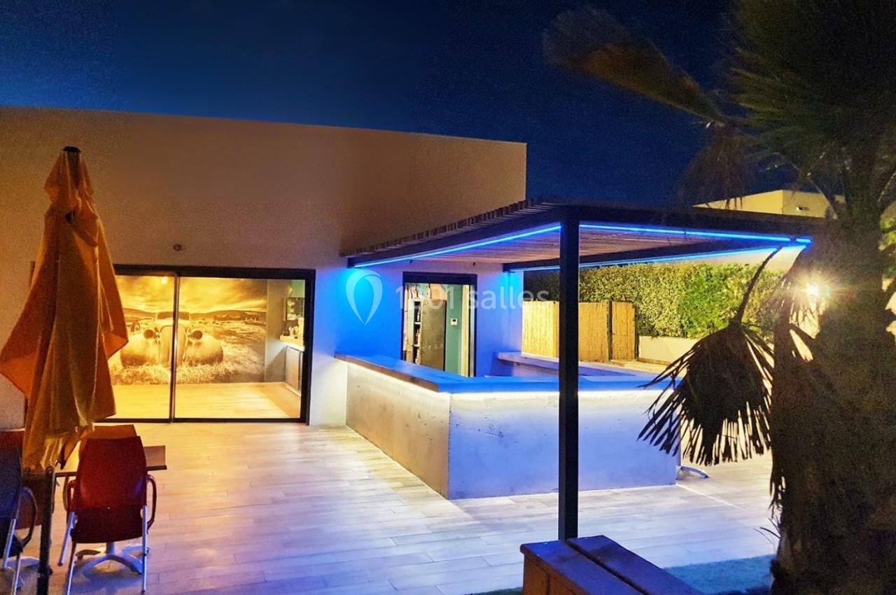 Terrasse moderne éclairée de nuit avec piscine, parasol jaune, palmiers et baie vitrée montrant un décor mural.