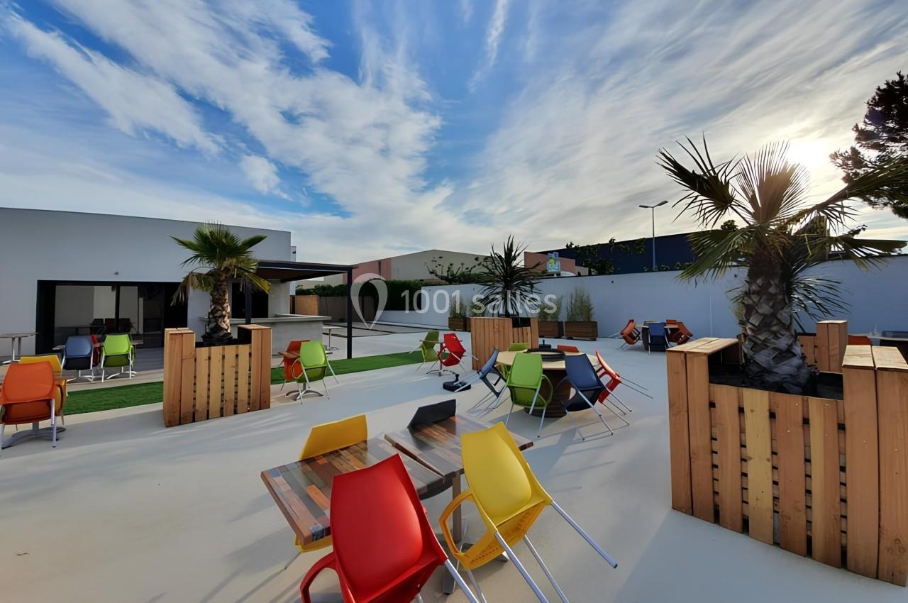 Terrasse extérieure avec tables en bois, chaises colorées et palmiers, sous un ciel partiellement nuageux.