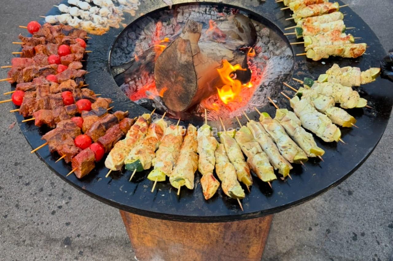 Brochettes variées grillant sur un barbecue circulaire avec un feu central, disposées en cercle.