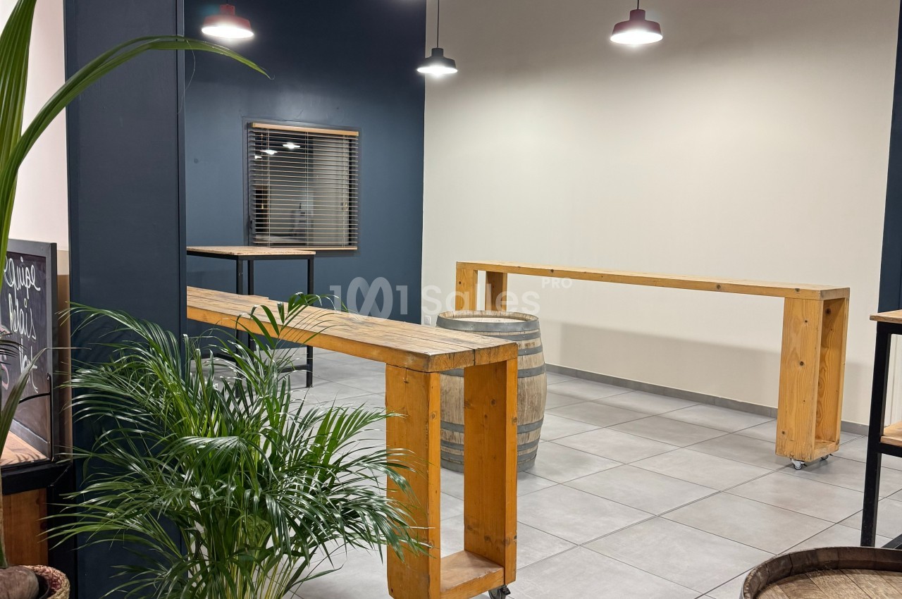 Salle moderne avec tables hautes en bois, tonneaux décoratifs, plantes vertes et éclairage suspendu.