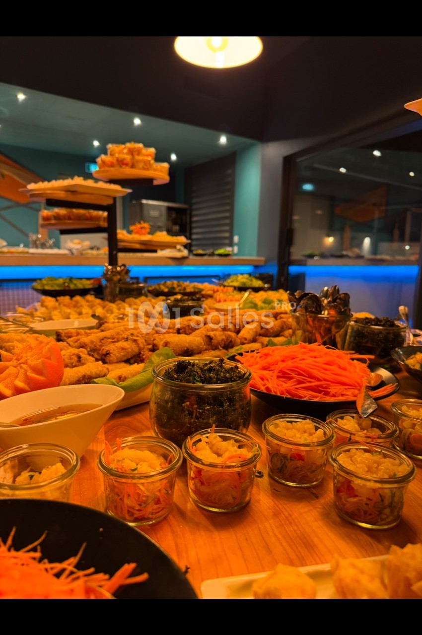 Buffet varié avec plats colorés, verrines, légumes et autres mets disposés sur une table en bois dans un cadre lumineux.