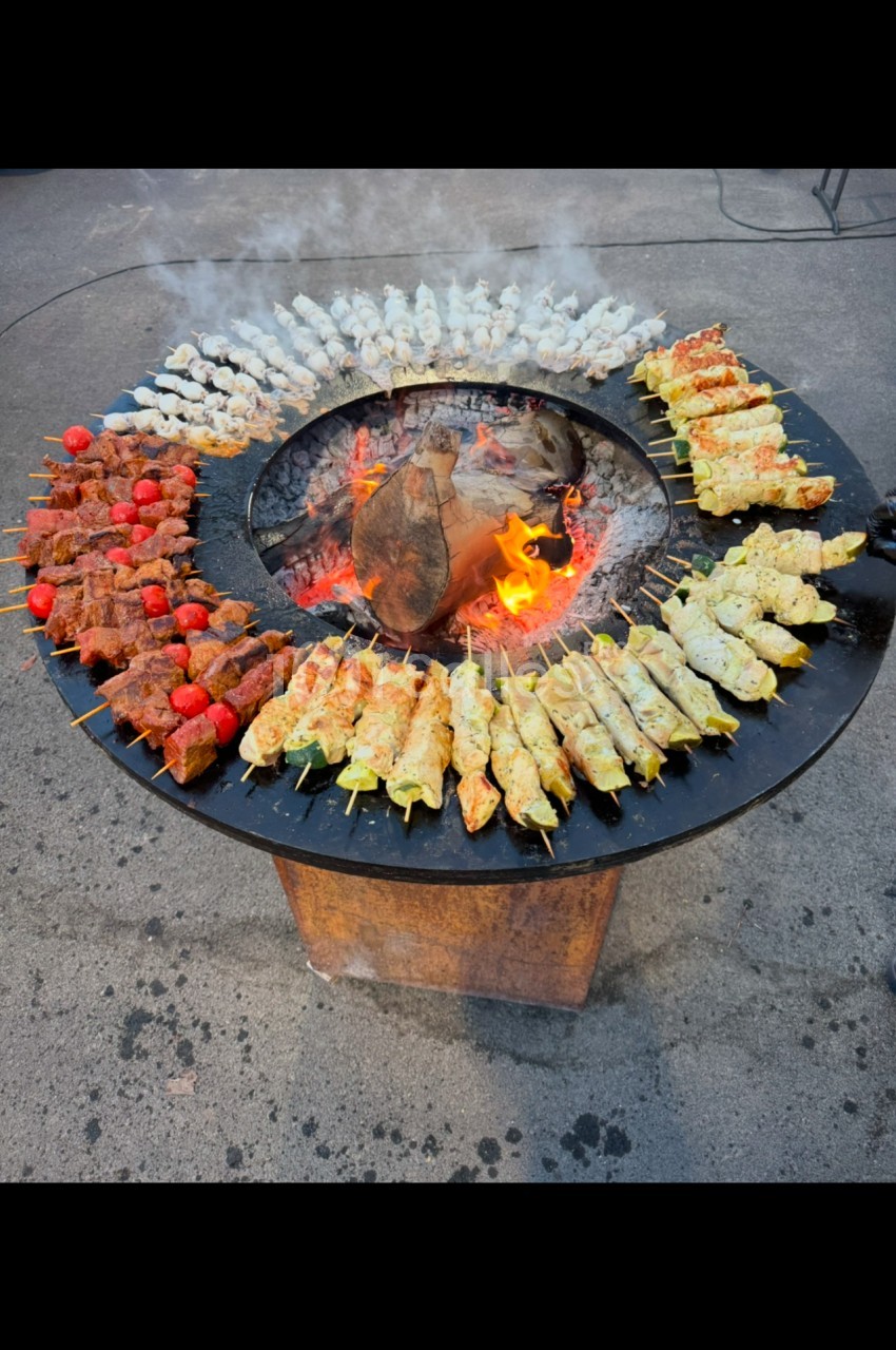 Brochettes variées grillant sur un barbecue circulaire avec un feu central, disposées en cercle.