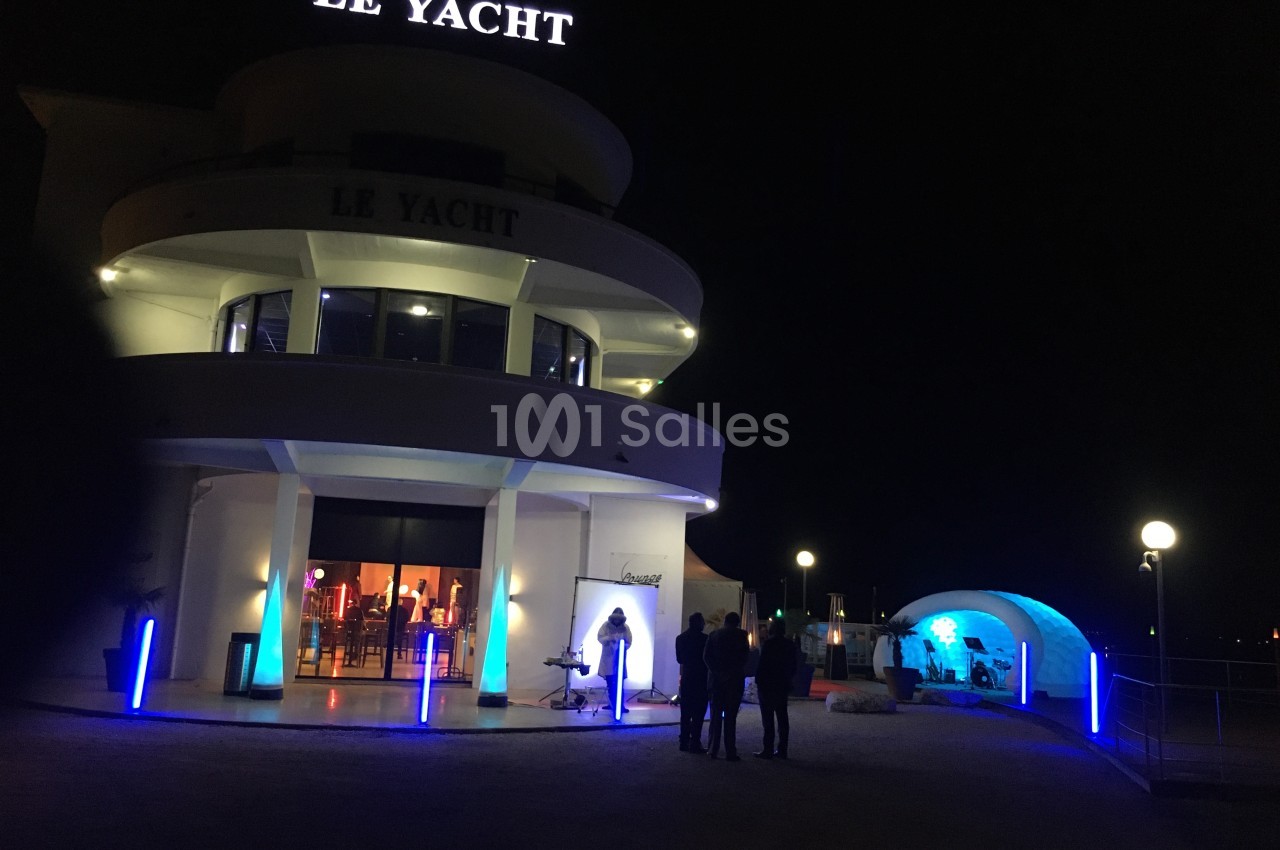Façade éclairée d'un bâtiment nommé ’Le Yacht’ de nuit, avec des lumières bleues et un espace extérieur aménagé.