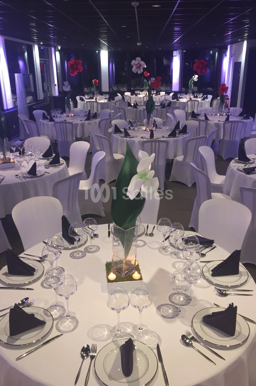 Salle de réception décorée avec des tables rondes, nappes blanches, serviettes noires et centres de table floraux.
