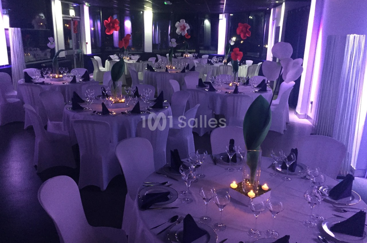 Salle de réception décorée avec des tables rondes, nappes blanches, serviettes pliées et éclairage tamisé violet.