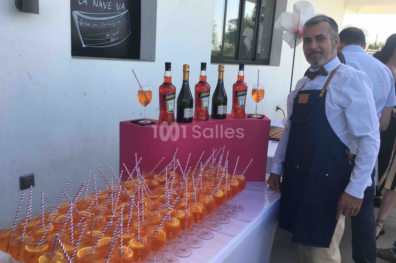Un serveur en tablier devant une table garnie de cocktails orange et de bouteilles d'alcool sur un présentoir rose.
