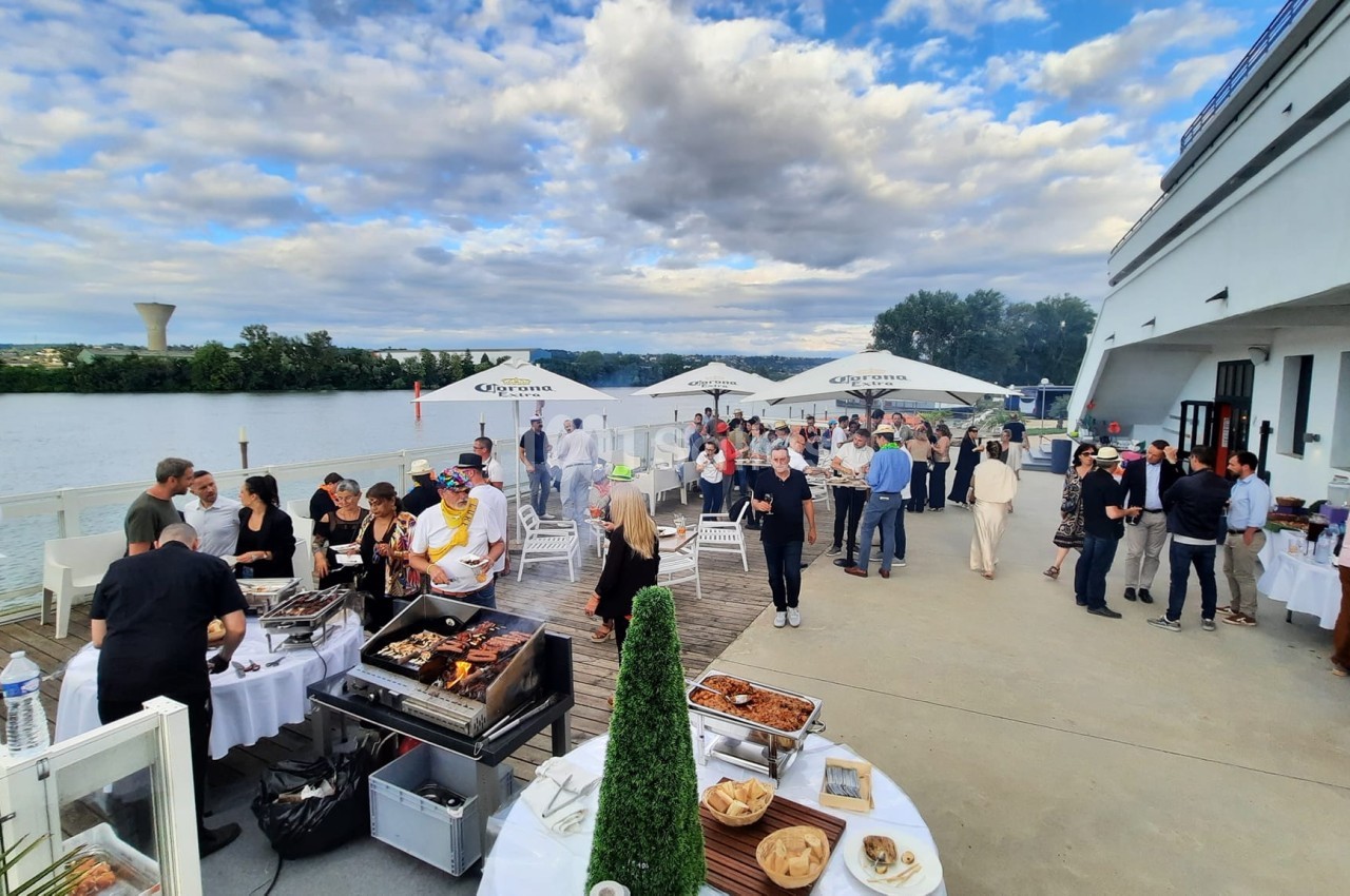 Personnes réunies sur une terrasse au bord d'un fleuve, avec des stands de nourriture et un ciel partiellement nuageux.