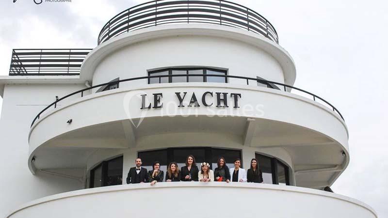 Façade blanche d'un bâtiment arrondi nommé ’Le Yacht’, avec plusieurs personnes souriantes sur un balcon.