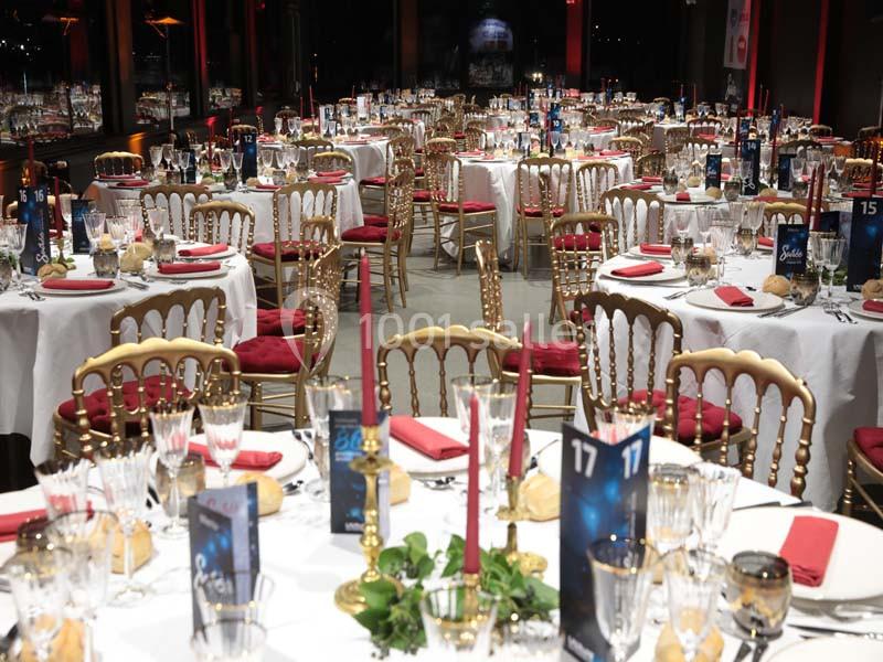 Salle de réception élégamment décorée avec des tables rondes, nappes blanches, chaises dorées et bougies rouges.