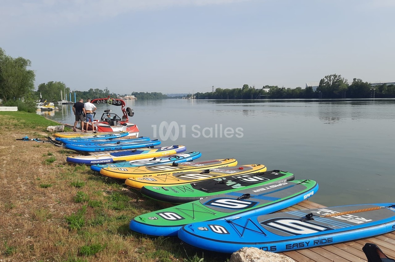 Plusieurs planches de paddle alignées au bord d'une rivière calme, avec quelques personnes et un bateau à moteur à proximité.