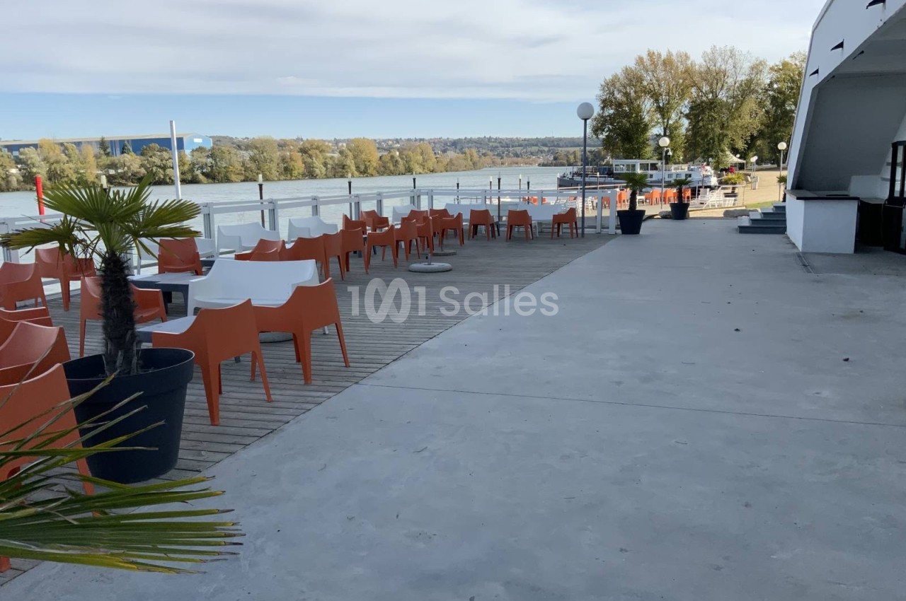 Terrasse avec chaises orange et tables blanches en bord de rivière, entourée de végétation et d'un ciel dégagé.