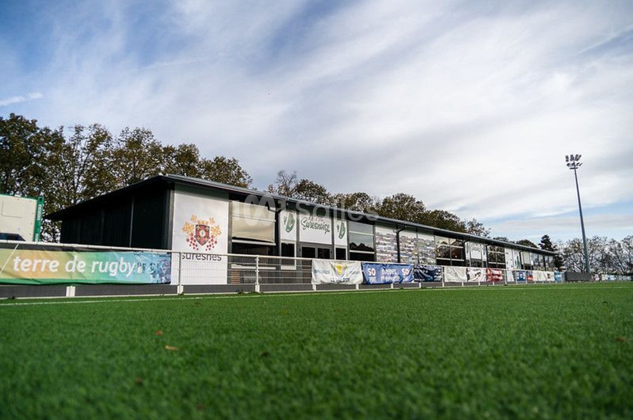 Terrain de rugby avec pelouse synthétique, bordé par un bâtiment et des panneaux publicitaires sous un ciel dégagé.