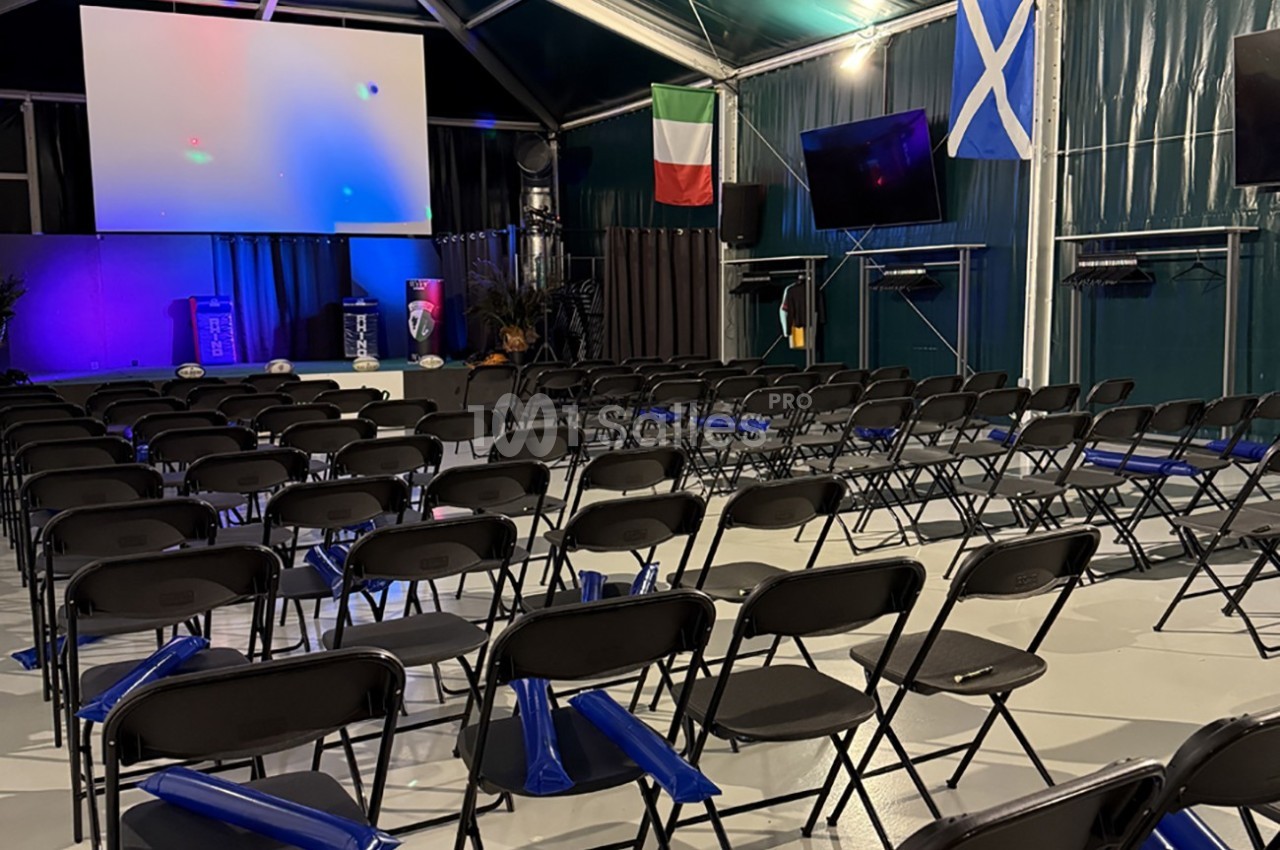 Salle de conférence vide avec des rangées de chaises noires, un écran géant et des drapeaux italien et écossais accrochés.