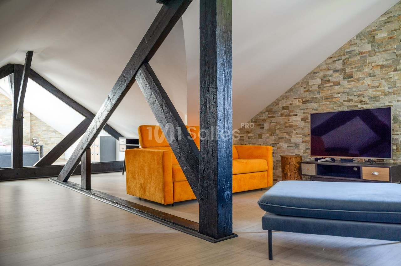 Salon mansardé avec poutres apparentes, canapé orange, mur en pierres et télévision sur meuble en bois.