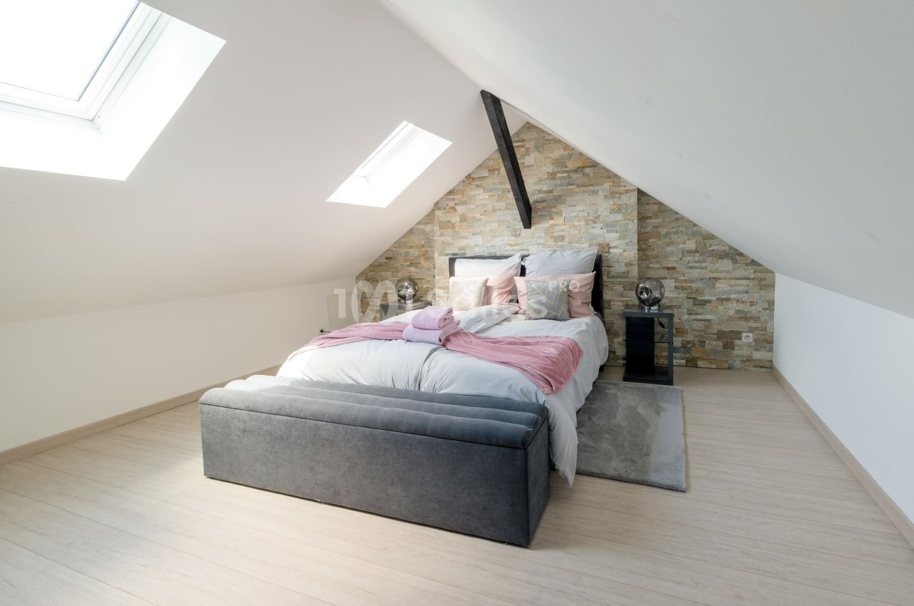 Chambre mansardée avec lit double, mur en pierre, lumière naturelle provenant de deux fenêtres de toit.