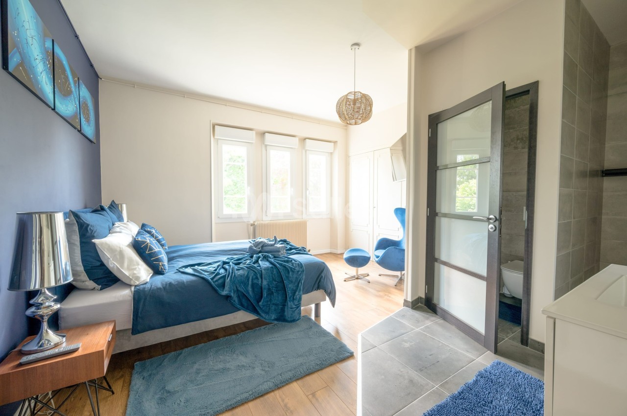 Chambre lumineuse avec lit double, draps bleus, murs blancs et bleus, fauteuil moderne et salle de bain attenante.