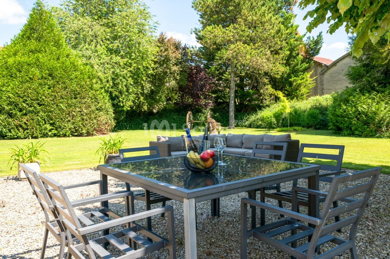 Table de jardin en métal avec chaises, décorée d'un panier de fruits, sur une terrasse en gravier entourée de verdure.