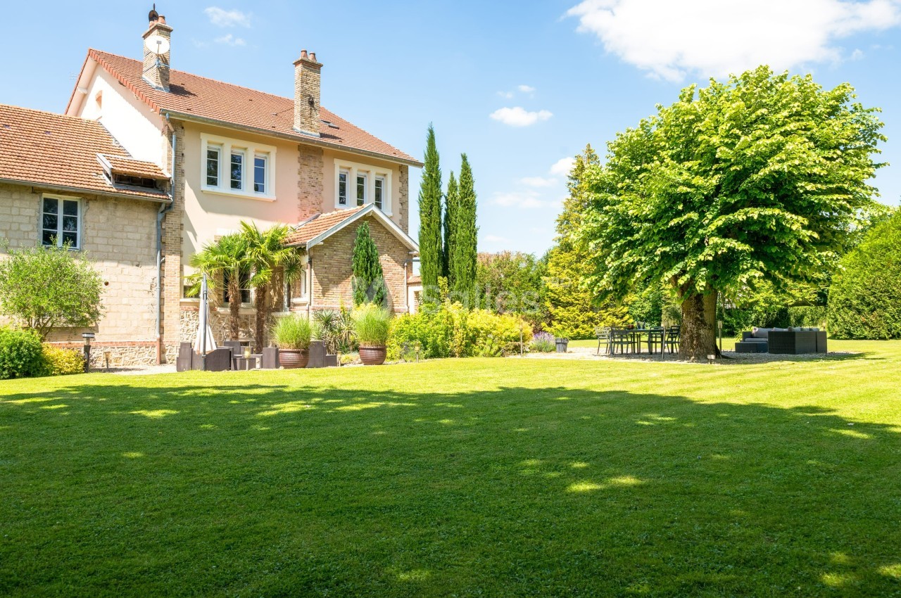 Jardin verdoyant avec pelouse, arbre ombragé, mobilier d'extérieur et maison en pierre sous un ciel bleu.