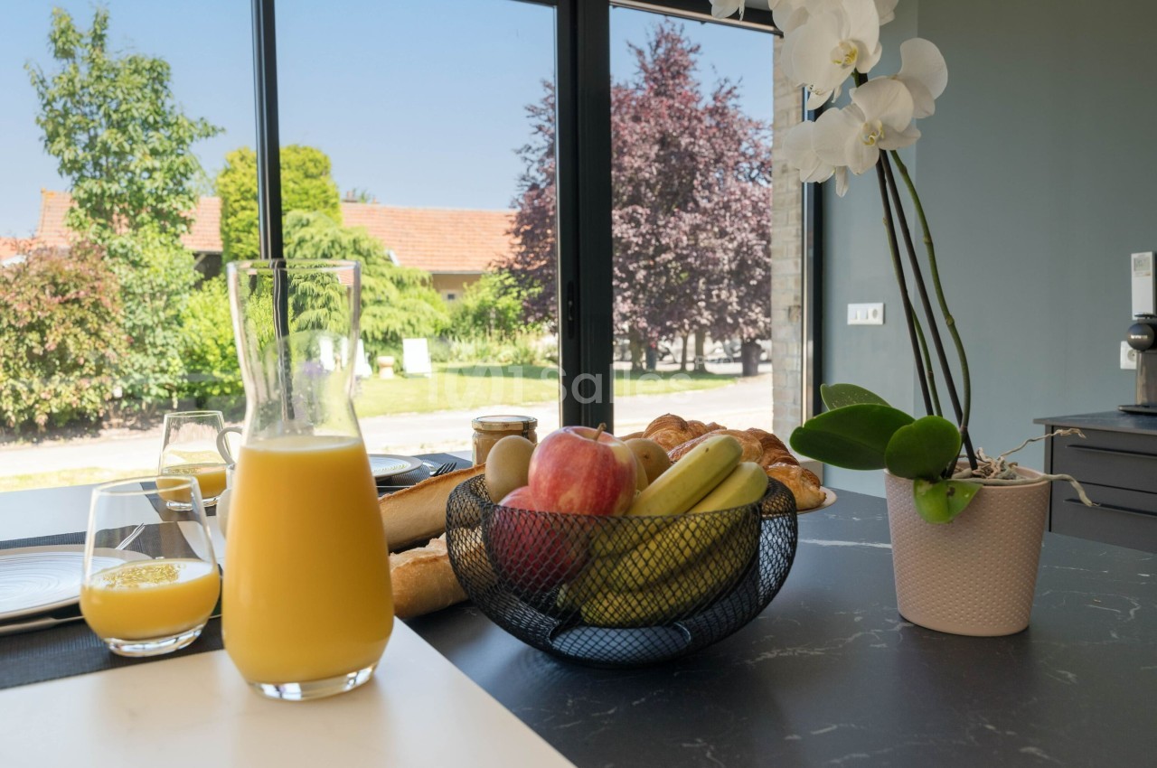 Petit-déjeuner sur une table avec jus d'orange, corbeille de fruits, viennoiseries et une orchidée en arrière-plan lumineux.
