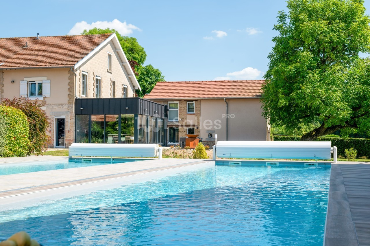 Piscine extérieure avec volets roulants, entourée de maisons en pierre et d'arbres sous un ciel bleu.