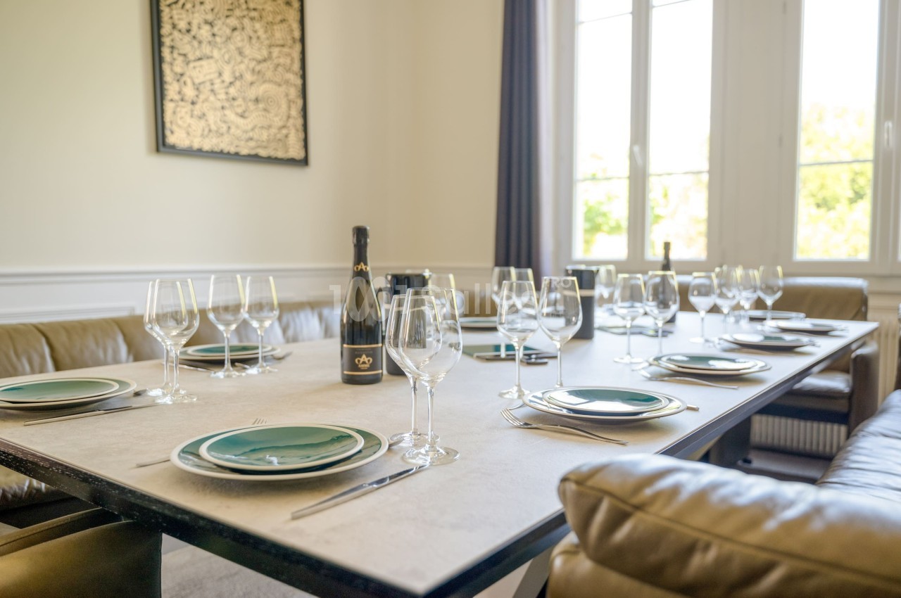 Table dressée avec des assiettes, verres à vin et une bouteille de champagne dans une salle lumineuse avec canapé en cuir.
