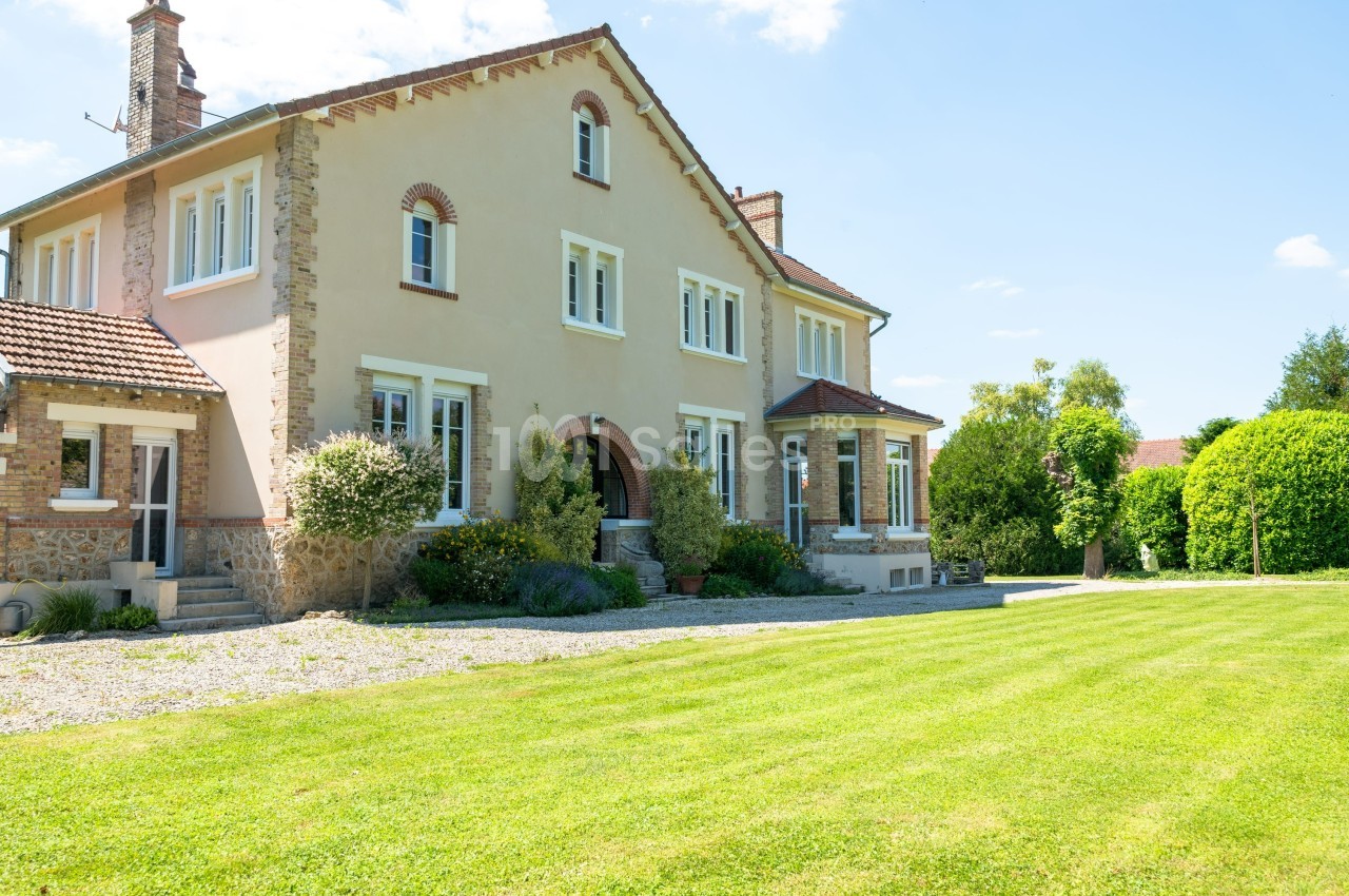 Grande maison en pierre et crépi beige avec jardin verdoyant et allée gravillonnée sous un ciel dégagé.