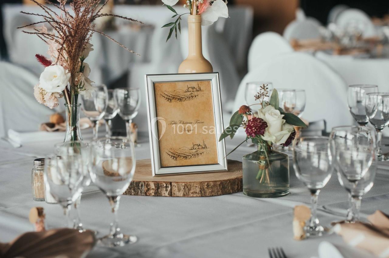 Table décorée pour un événement avec nappes blanches, verres, couverts, et centre de table floral avec cadre en bois.