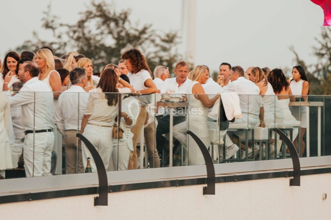 Groupe de personnes en tenue blanche réunies sur une terrasse en hauteur, avec des arbres et une éolienne en arrière-plan.
