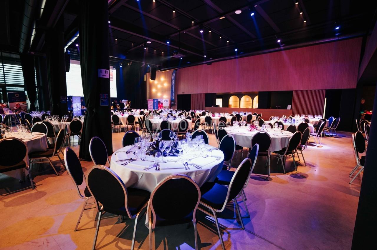 Salle de réception avec tables rondes dressées, chaises noires et éclairage tamisé dans un espace moderne.