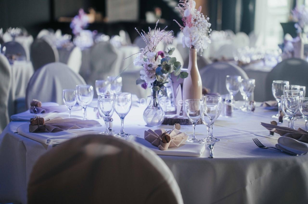 Table ronde décorée pour un événement, avec nappes blanches, verres, serviettes pliées et bouquets de fleurs.