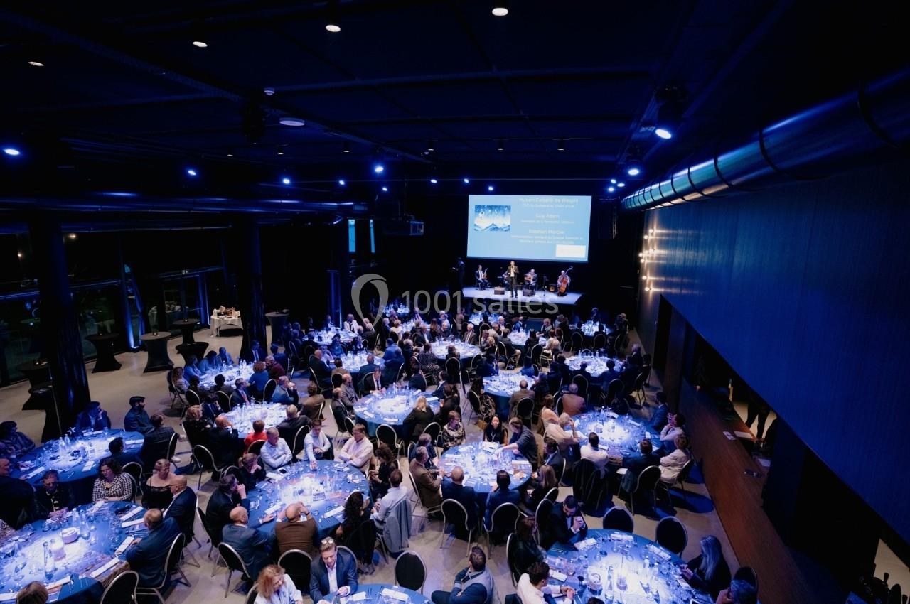 Salle de réception sombre avec plusieurs tables rondes illuminées, occupées par des convives lors d'un événement.