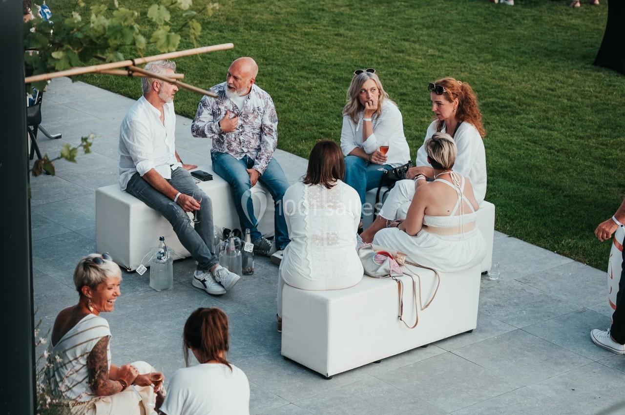 Un groupe de personnes discute assises sur des bancs blancs en extérieur, sur une terrasse près d'une pelouse.