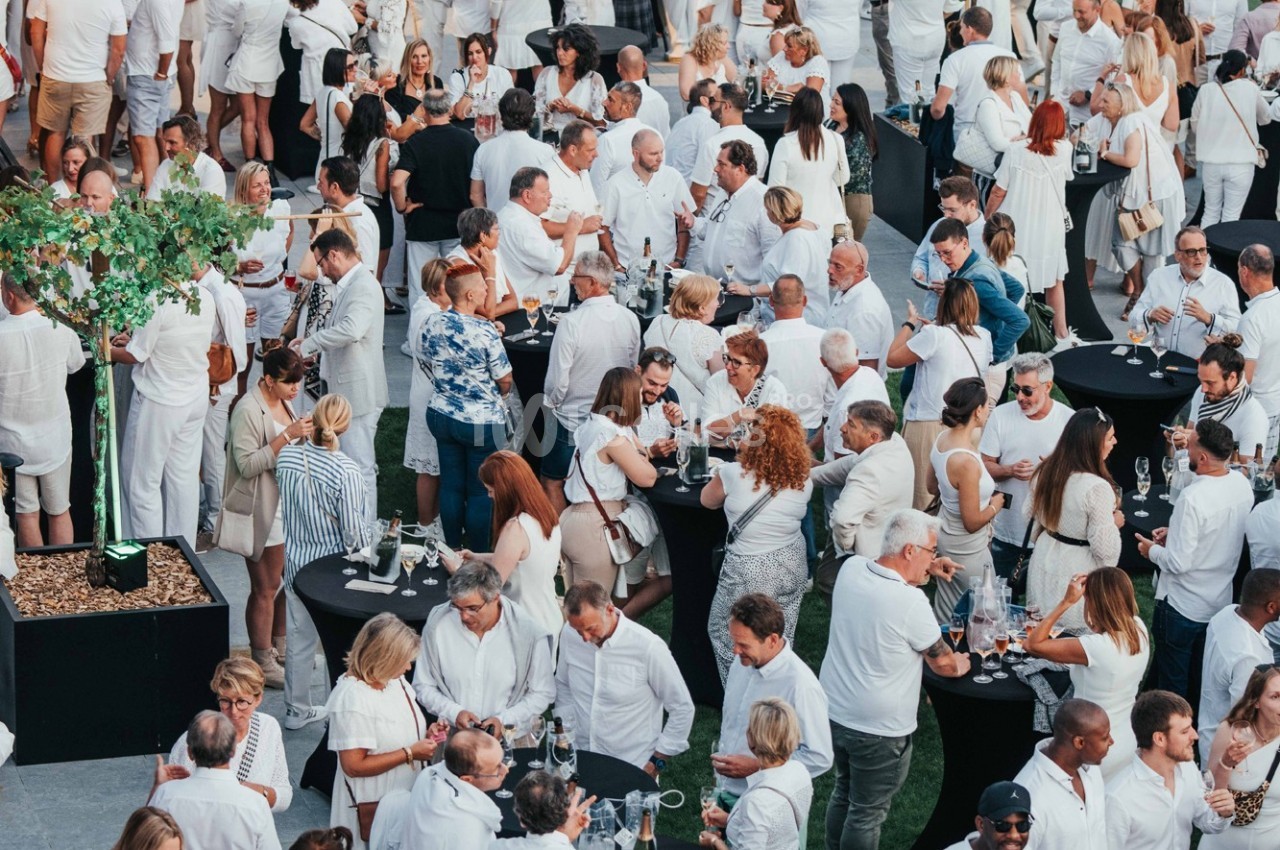 Groupe de personnes en tenue blanche rassemblées autour de tables noires lors d'un événement en plein air.