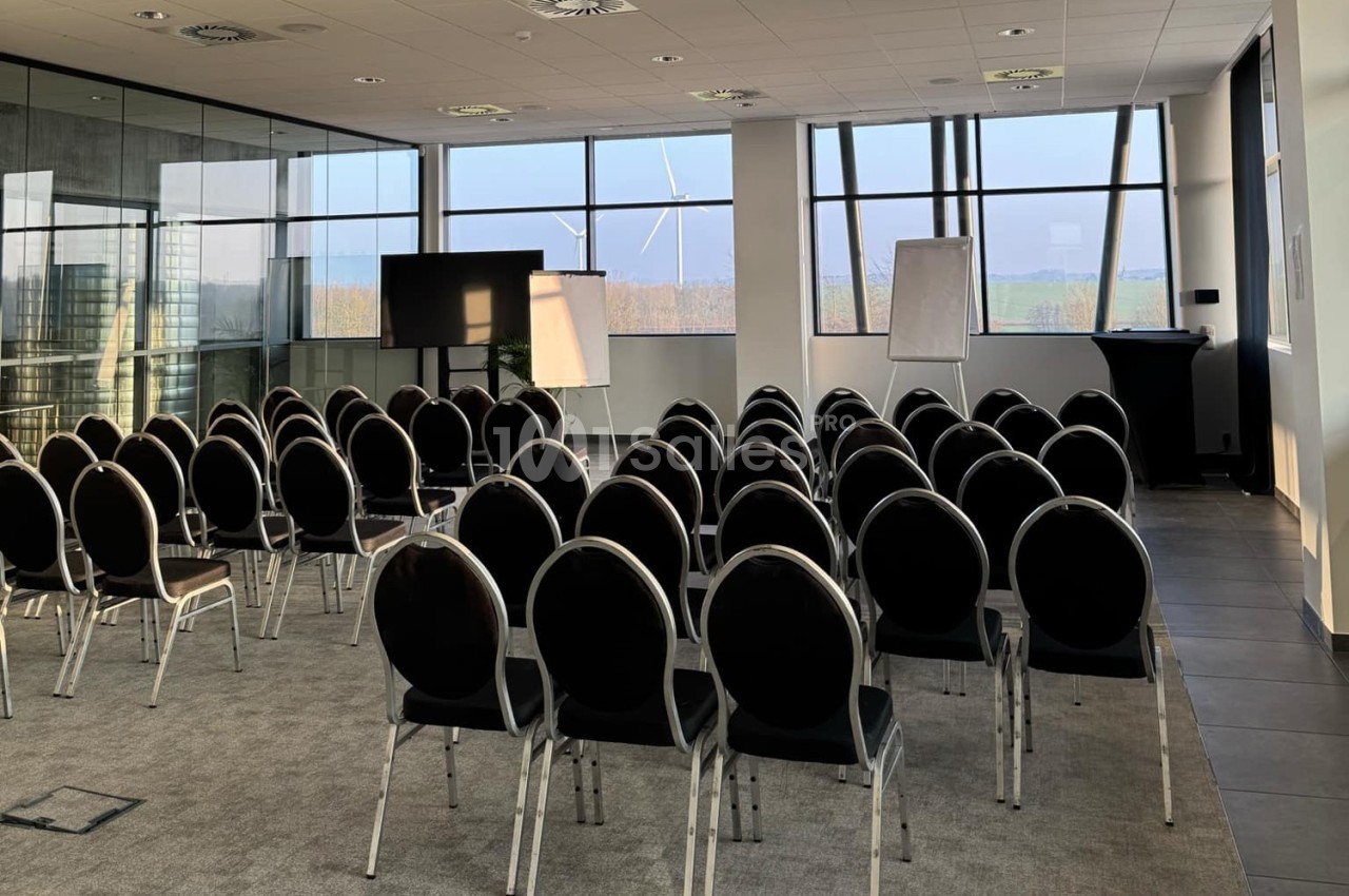 Salle de conférence lumineuse avec des rangées de chaises noires, un écran, des tableaux blancs et de grandes fenêtres.