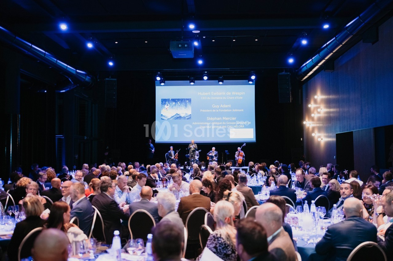 Salle de réception avec des invités assis à des tables, une scène avec des musiciens et un écran projetant des informations.
