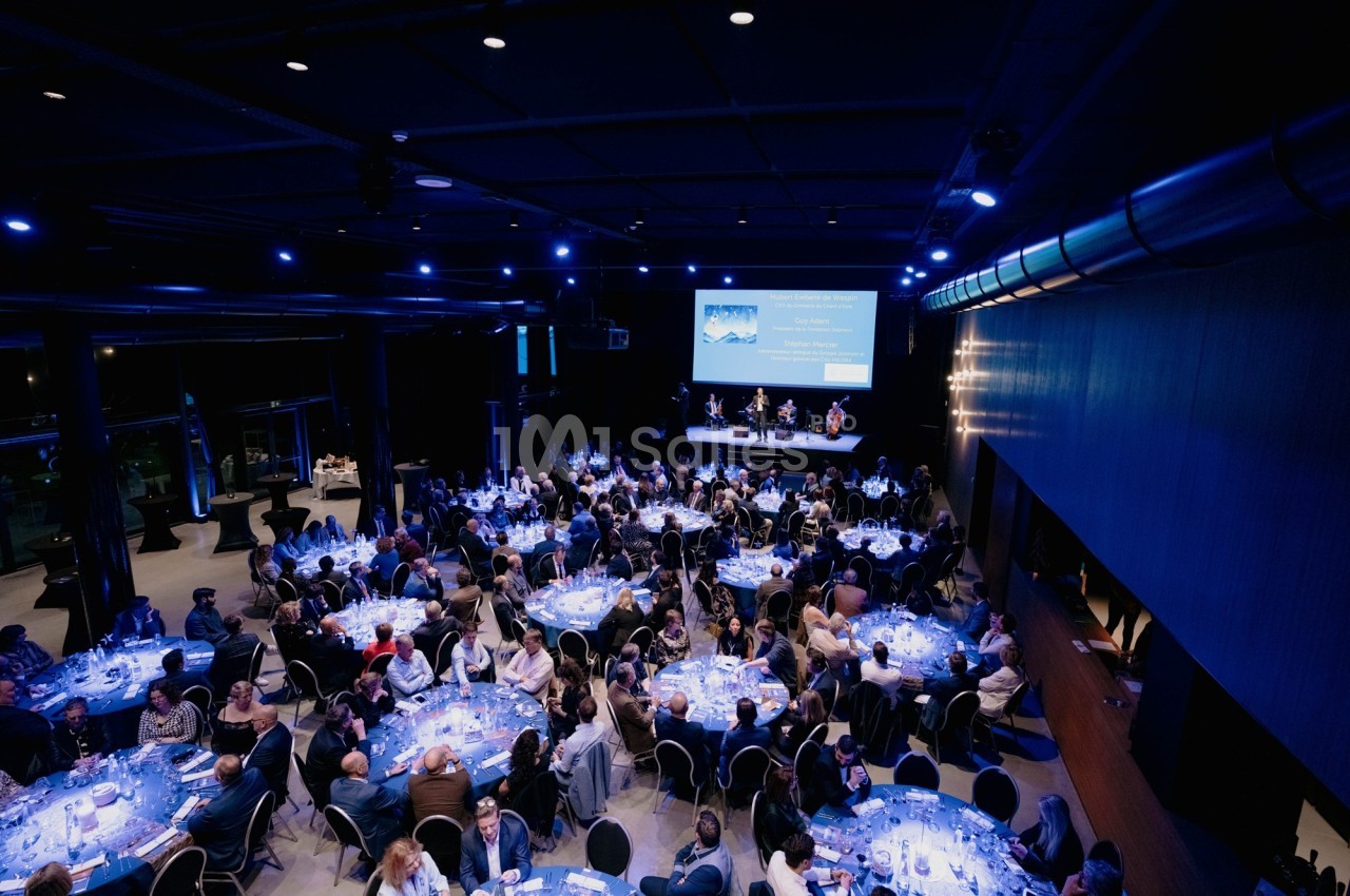 Salle de réception sombre avec plusieurs tables rondes illuminées, occupées par des convives lors d'un événement.