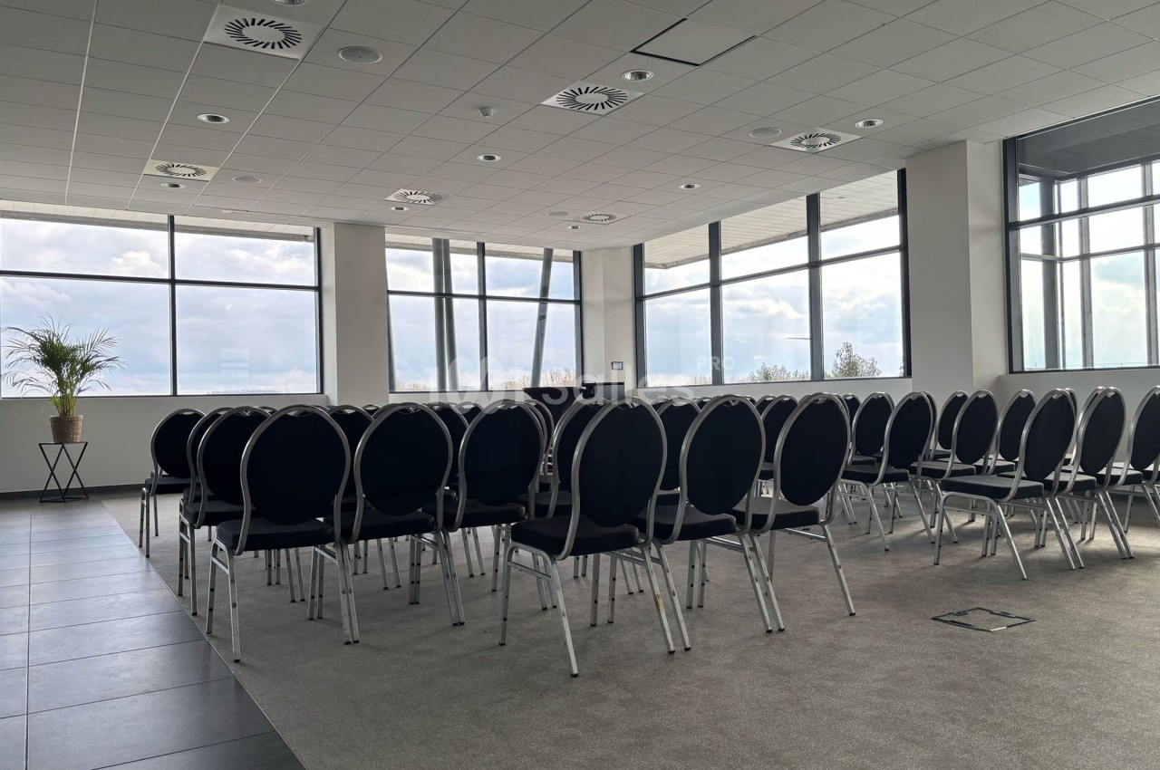 Salle de conférence lumineuse avec des rangées de chaises noires alignées face à des fenêtres panoramiques.