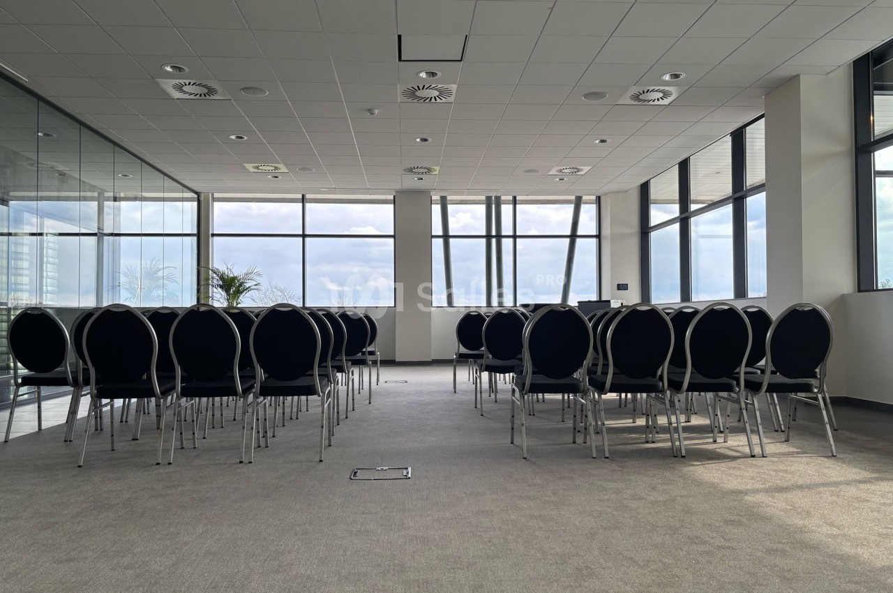 Salle de réunion lumineuse avec des rangées de chaises noires alignées face à des baies vitrées.
