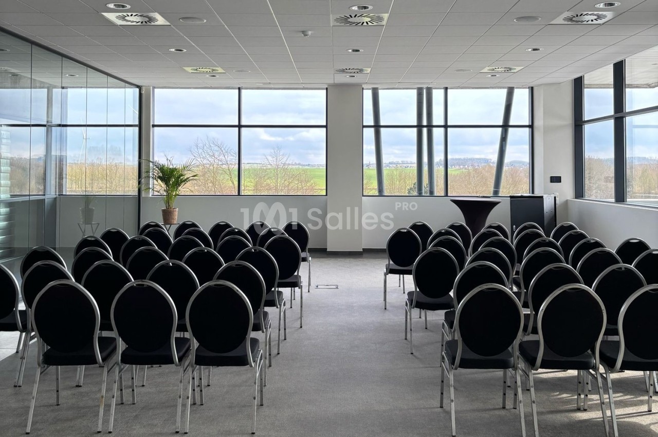 Salle de conférence lumineuse avec des rangées de chaises noires alignées face à une baie vitrée donnant sur un paysage.