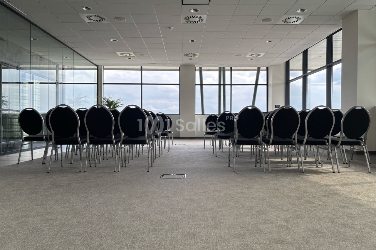 Salle de conférence lumineuse avec des rangées de chaises noires alignées face à une grande baie vitrée.