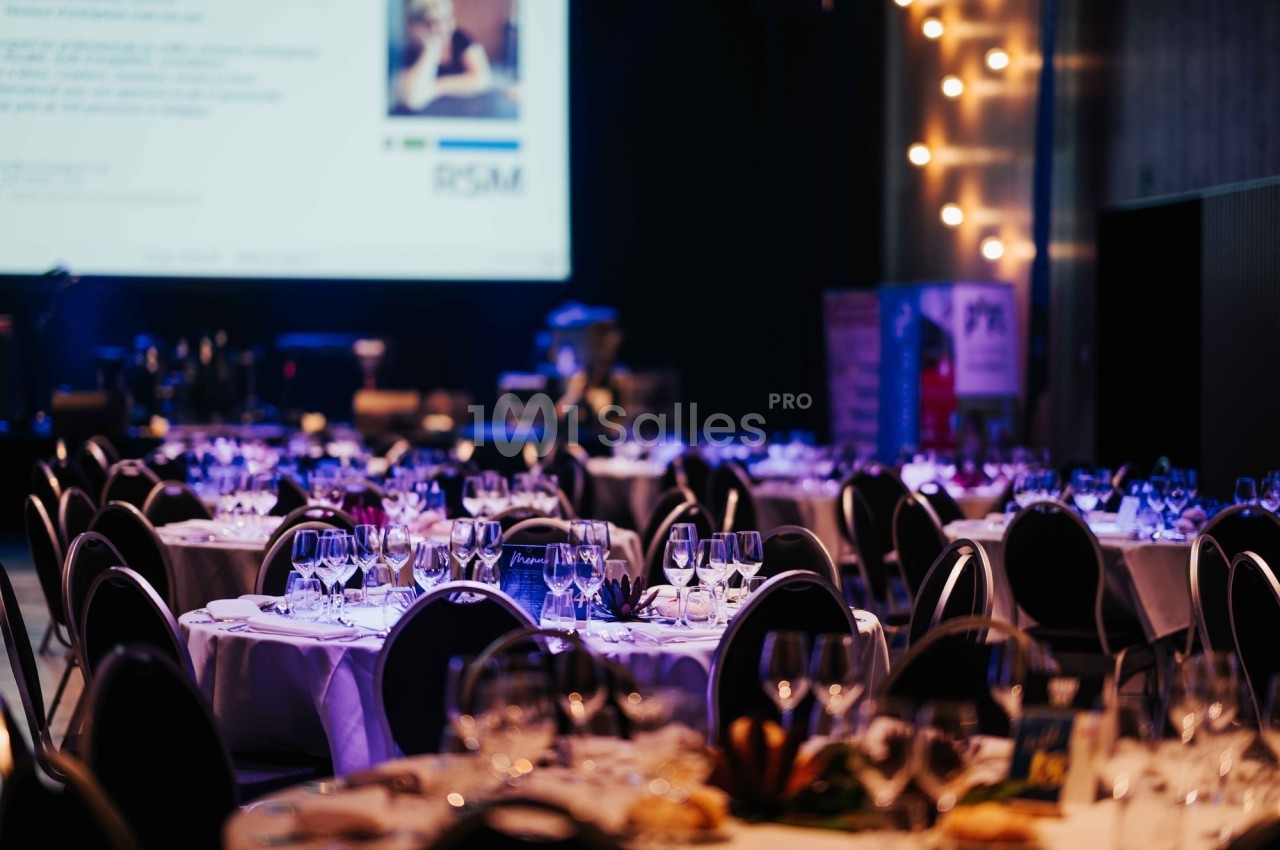 Salle de réception avec tables rondes dressées, verres et couverts, éclairage tamisé et écran en arrière-plan.