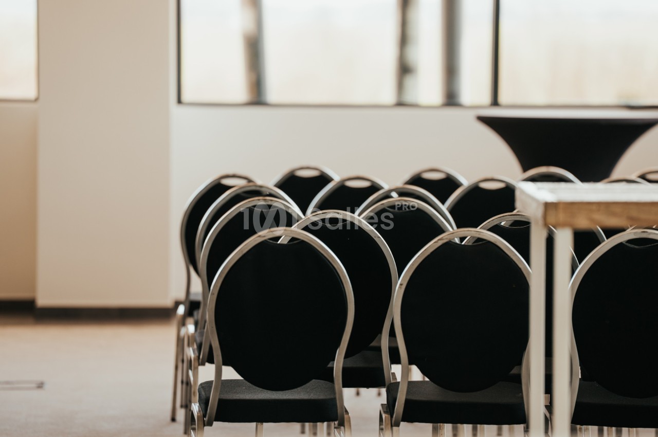 Rangées de chaises noires alignées dans une salle lumineuse avec de grandes fenêtres en arrière-plan.