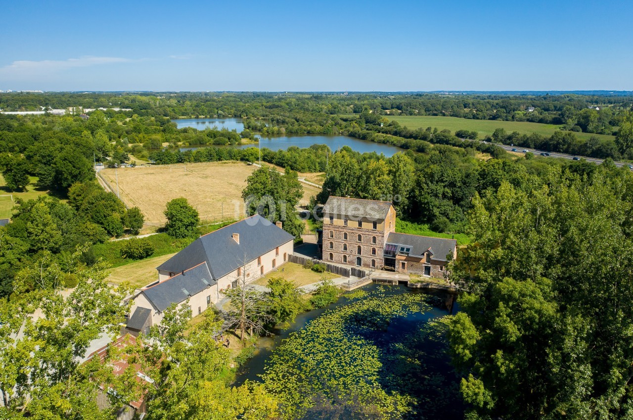 Vue aérienne d'un moulin restauré avec étang, entouré de prairies, de forêts et de plans d'eau sous un ciel dégagé.