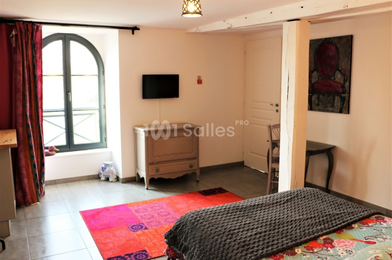 Chambre lumineuse avec lit, télévision murale, fenêtre cintrée, mobilier ancien et décoration colorée.