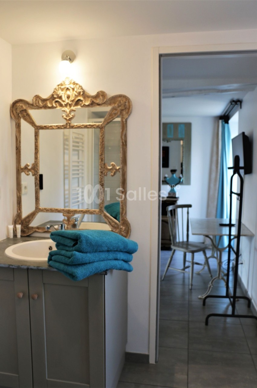 Salle de bain avec un lavabo, un miroir doré orné, des serviettes bleues pliées et vue sur une pièce adjacente.